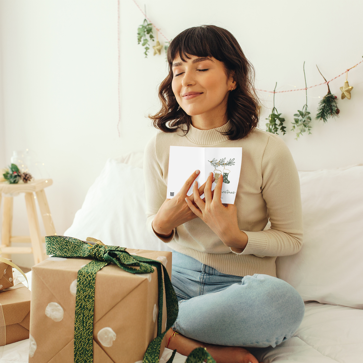 A woman sits on a white sofa in a bright, cozy room, holding a small white card with a green illustration. A wrapped gift box with a green ribbon is in the foreground, and holiday decorations hang on the wall behind her.