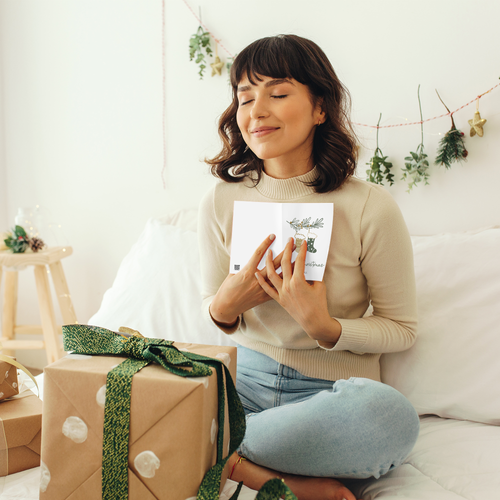 A woman sits on a white sofa in a bright, cozy room, holding a small white card with a green illustration. A wrapped gift box with a green ribbon is in the foreground, and holiday decorations hang on the wall behind her.